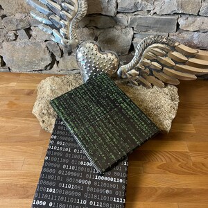 以下が含まれることがあります： 緑と白のバイナリコードのパターンが施された黒いノートが2冊。ノートは重ねられています。背景には羽根のついた銀色のハートがあります。