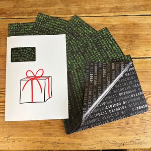 以下が含まれることがあります： 赤いリボンと弓が付いた白いギフトボックス。ボックスは、バイナリコードの黒と緑のパターンが施された白い背景の上にあります。