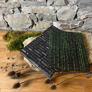 以下が含まれることがあります： 緑色のバイナリコードのパターンが施された黒いノートが2冊。ノートは重ねて木製の表面に置かれています。