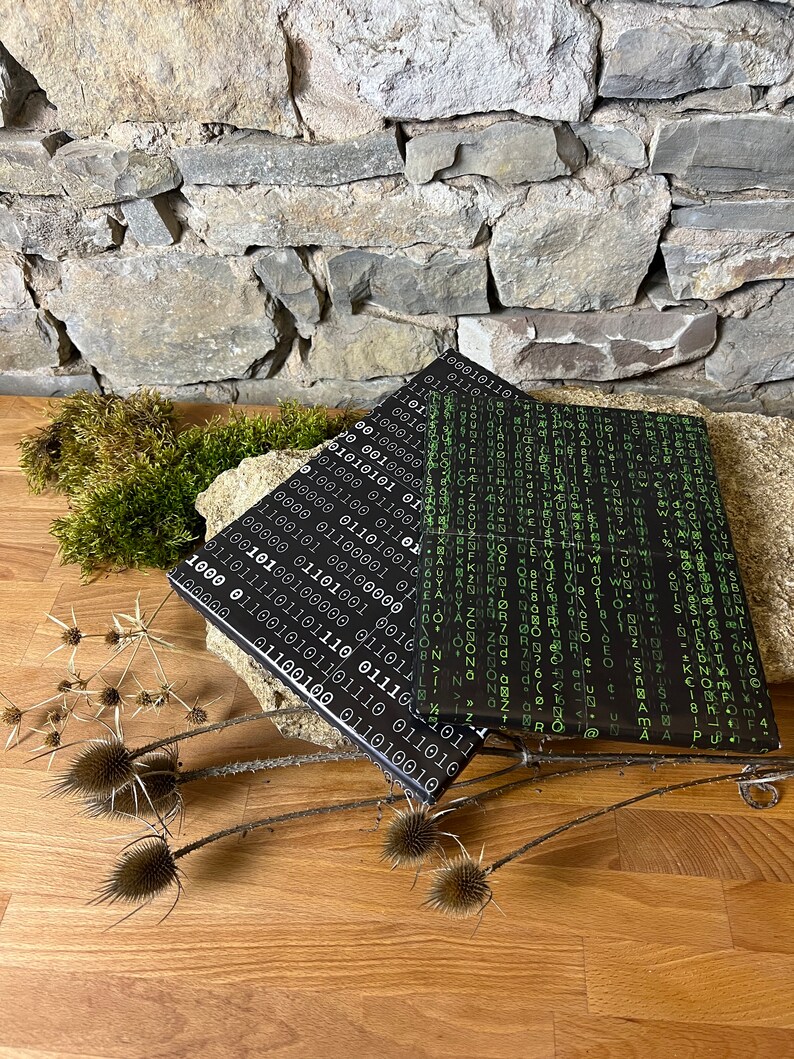 以下が含まれることがあります： 緑色のバイナリコードのパターンが施された黒いノートが2冊。ノートは重ねて木製の表面に置かれています。