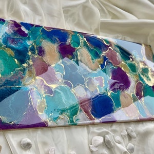 Puede incluir: Obra de arte abstracta con acabado brillante. La obra presenta una mezcla de colores turquesa, azul, morado y dorado. La pieza rectangular est&aacute; montada sobre un marco de madera, creando una est&eacute;tica moderna y art&iacute;stica.
