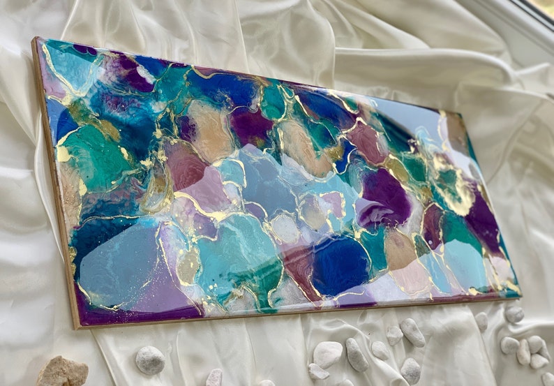 Puede incluir: Obra de arte abstracta con acabado brillante. La obra presenta una mezcla de colores turquesa, azul, morado y dorado. La pieza rectangular est&aacute; montada sobre un marco de madera, creando una est&eacute;tica moderna y art&iacute;stica.