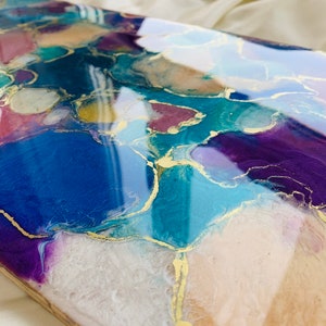 Puede incluir: Pieza de arte abstracto con un acabado brillante. La obra presenta una mezcla de colores, incluyendo azul, morado, verde azulado y dorado, creando un efecto marmolado. La pieza es rectangular y parece estar hecha de resina o un material similar.