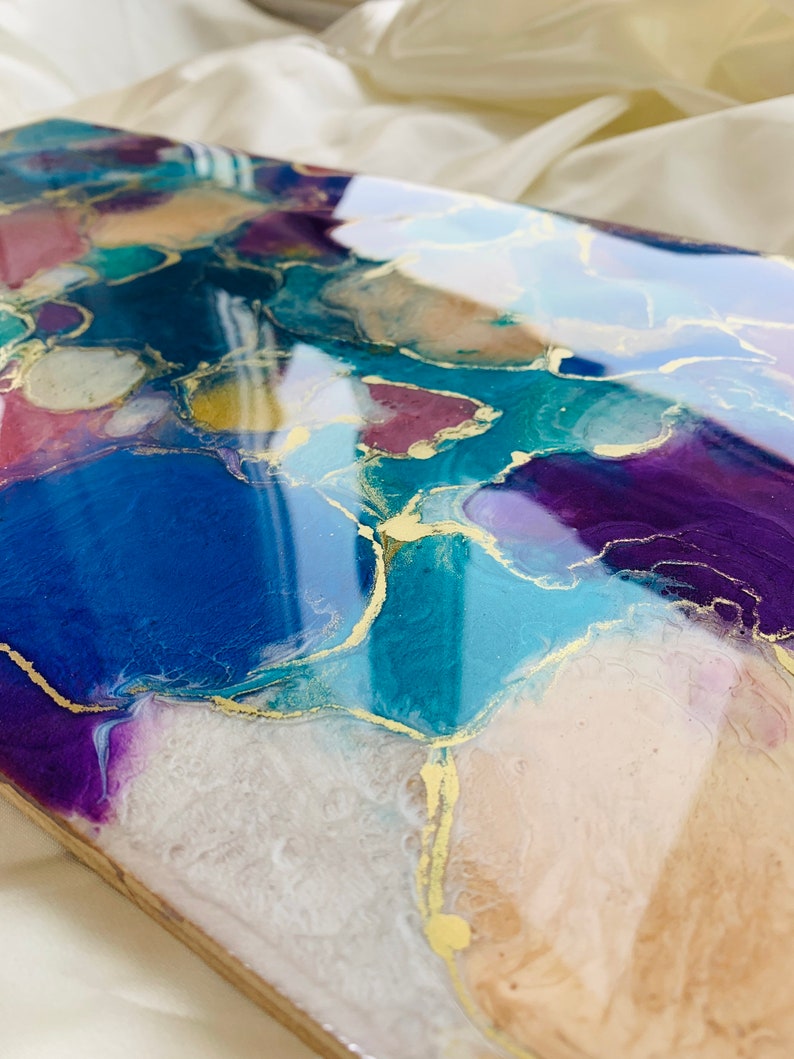 Puede incluir: Pieza de arte abstracto con un acabado brillante. La obra presenta una mezcla de colores, incluyendo azul, morado, verde azulado y dorado, creando un efecto marmolado. La pieza es rectangular y parece estar hecha de resina o un material similar.