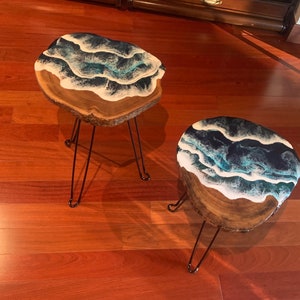 Könnte beinhalten: Zwei handgefertigte Beistelltische aus Holz mit Epoxidharzplatten. Die Oberflächen weisen ein blau-weißes Wellenmuster auf. Jeder Tisch hat eine natürliche Holzkante und schwarze Haarnadelbeine. Ein einzigartiger Einrichtungsgegenstand.