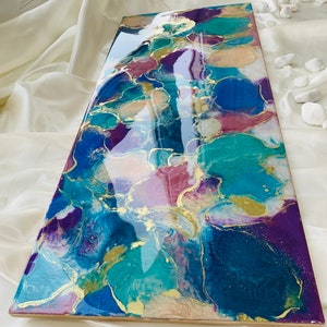 Puede incluir: Pieza de arte abstracto con acabado brillante. La obra presenta una mezcla de azules, turquesas, morados y acentos dorados. La pieza rectangular se exhibe sobre una superficie blanca, mostrando los colores y texturas vibrantes.