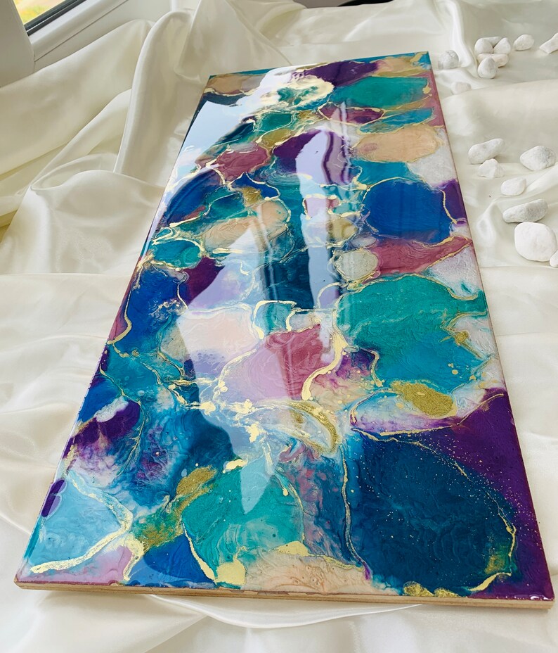 Puede incluir: Pieza de arte abstracto con acabado brillante. La obra presenta una mezcla de azules, turquesas, morados y acentos dorados. La pieza rectangular se exhibe sobre una superficie blanca, mostrando los colores y texturas vibrantes.