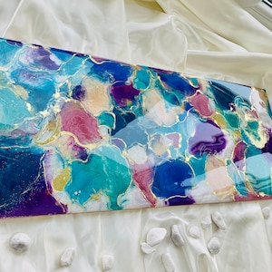 Puede incluir: Obra de arte abstracta con acabado brillante. La obra presenta una mezcla de azules, turquesas, p&uacute;rpuras y rosas, acentuada con pan de oro. La pieza rectangular se exhibe sobre una superficie blanca texturizada.