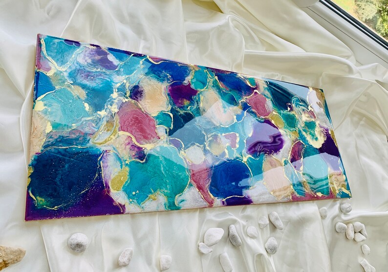 Puede incluir: Obra de arte abstracta con acabado brillante. La obra presenta una mezcla de azules, turquesas, p&uacute;rpuras y rosas, acentuada con pan de oro. La pieza rectangular se exhibe sobre una superficie blanca texturizada.