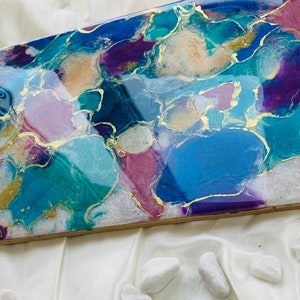 Puede incluir: Pieza de arte abstracto de resina con forma rectangular. La obra presenta una mezcla de azules, morados y acentos dorados. Los colores se arremolinan, creando un dise&ntilde;o &uacute;nico y llamativo. La pieza est&aacute; sobre un fondo blanco.