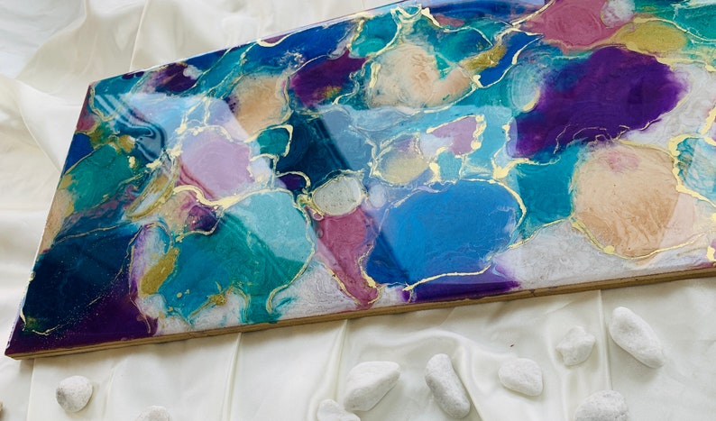 Puede incluir: Pieza de arte abstracto de resina con forma rectangular. La obra presenta una mezcla de azules, morados y acentos dorados. Los colores se arremolinan, creando un dise&ntilde;o &uacute;nico y llamativo. La pieza est&aacute; sobre un fondo blanco.