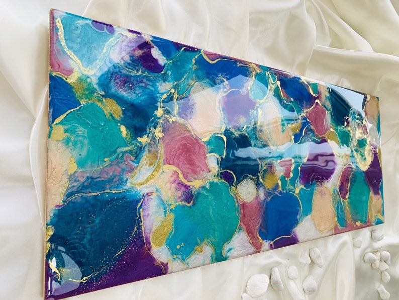 Puede incluir: Obra de arte abstracta de resina con acabado brillante. La obra presenta una mezcla de colores turquesa, azul, morado y dorado. El dise&ntilde;o tiene una apariencia fluida y org&aacute;nica, con acentos dorados que crean un efecto marmoleado. La pieza es rectangular.