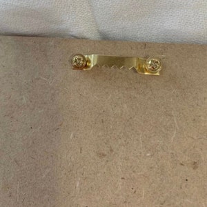 Puede incluir: Primer plano de la parte posterior de un marco de fotos de madera. Un colgador de dientes de sierra de color dorado est&aacute; sujeto con dos tornillos. El marco est&aacute; hecho de un material marr&oacute;n claro y texturizado.