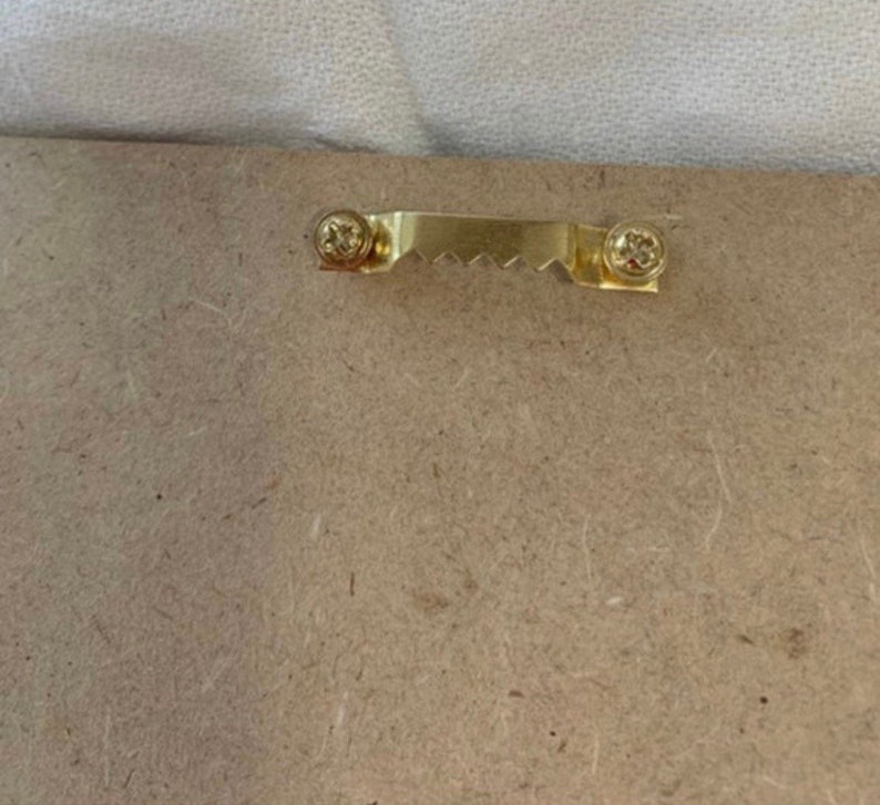 Puede incluir: Primer plano de la parte posterior de un marco de fotos de madera. Un colgador de dientes de sierra de color dorado est&aacute; sujeto con dos tornillos. El marco est&aacute; hecho de un material marr&oacute;n claro y texturizado.