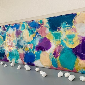 Puede incluir: Obra de arte abstracta con una vibrante mezcla de colores turquesa, azul, morado y dorado. La obra presenta un dise&ntilde;o org&aacute;nico y fluido con un acabado brillante. Guijarros blancos est&aacute;n colocados frente a la obra.