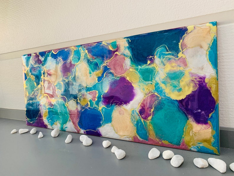 Puede incluir: Obra de arte abstracta con una vibrante mezcla de colores turquesa, azul, morado y dorado. La obra presenta un dise&ntilde;o org&aacute;nico y fluido con un acabado brillante. Guijarros blancos est&aacute;n colocados frente a la obra.
