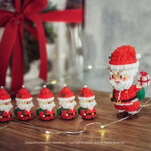 Può includere: Figure pixelate di Babbo Natale, con abiti e cappelli rossi, barbe bianche e regali. Le figure sono disposte su una superficie di legno. Lo sfondo è sfocato, con luci a catena che aggiungono un tocco festoso.