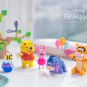 Puede incluir: Una colección de figuras de personajes pixelados y coloridos, que incluyen a Winnie the Pooh, Piglet, Eeyore y Tigger, junto con un árbol, una abeja y globos. También es visible el texto "Don't Worry Be Happy".
