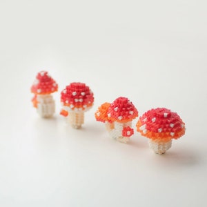 May include: Four small, handmade mushroom figurines. The mushroom caps are red and orange with white spots, and the stems are white. The mushrooms are arranged in a row, with varying sizes and details, creating a whimsical, decorative theme.