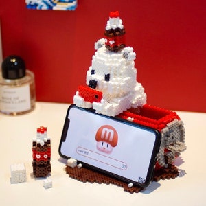 Puede incluir: Un oso polar blanco de pixel art sosteniendo una tableta roja, con un smartphone delante. El teléfono muestra un logotipo de hongo. Un pequeño cupcake de pixel art está en la cabeza del oso. La base es marrón.