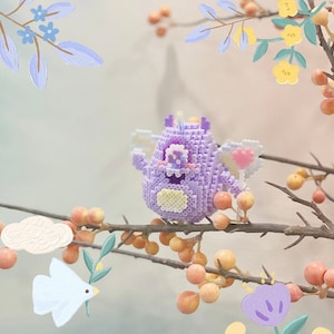 Puede incluir: Una criatura púrpura pixelada y caprichosa con alas en forma de corazón y una barriga amarilla. La figura hecha a mano está posada en una rama con bayas, rodeada de acentos florales pintados en una paleta de colores pastel suaves. Un artículo decorativo único.