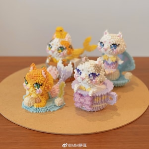 Puede incluir: Cuatro figuras de gatos coloridos hechas de pequeños bloques. Los gatos están en varias poses, con detalles como una taza de té, una cola de sirena y otros accesorios. La paleta de colores incluye naranja, blanco, amarillo y azul.