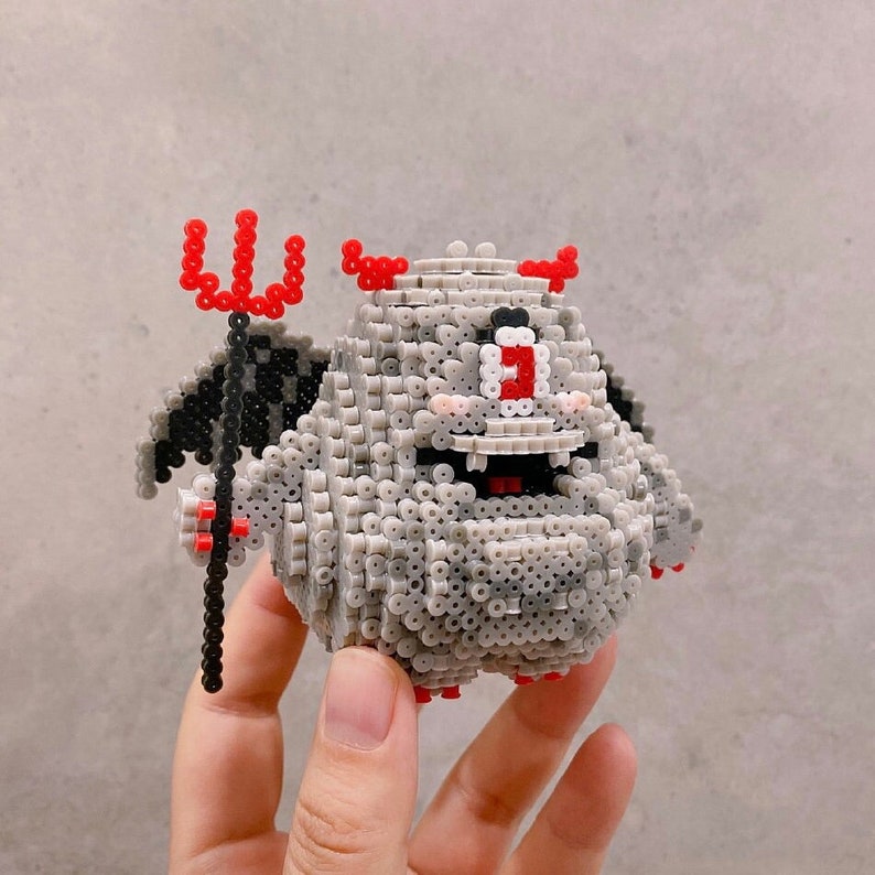 Pu&ograve; includere: Una figura di diavolo in pixel art grigio con corna rosse, un tridente rosso e ali nere. Il diavolo ha un ampio sorriso, un solo occhio e guance rosee. La figura &egrave; fatta di piccole perline rotonde.