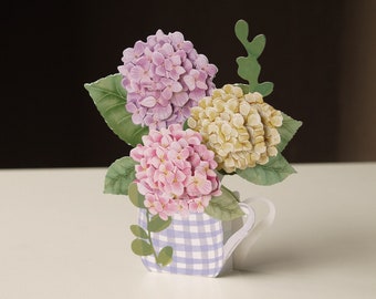 Peut inclure: Une carte pop-up 3D représentant un bouquet de trois hortensias dans un vase à carreaux bleus et blancs. Les hortensias sont roses, violets et jaunes.
