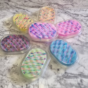 May include: Seven oval sponges in clear plastic containers. Each sponge features a unique colour scheme, including yellow, pink, blue, green, and purple, with streaks of other colours. The sponges are arranged in a circular pattern.