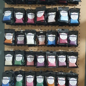May include: A wall display of small, clear plastic bags filled with colourful bath salts. The bags are hung on a pegboard with a variety of colours, including pink, blue, green, purple, and orange.