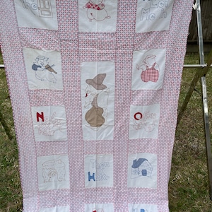 May include: A vintage quilt with a pink and white checkered pattern. The quilt is embroidered with scenes from the children's story "The Three Little Pigs". The text "Open the door", "And Let Me In", "No! by the hair", "Then huff", "I'll blow you're in!" and "And Jill Puff" are embroidered on the quilt.