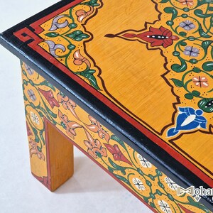 May include: A low, wooden table with a vibrant, hand-painted design. The tabletop is a warm yellow with intricate floral patterns in red, green, white, and blue. The edges are outlined in black and red. The table's legs are a natural wood color.