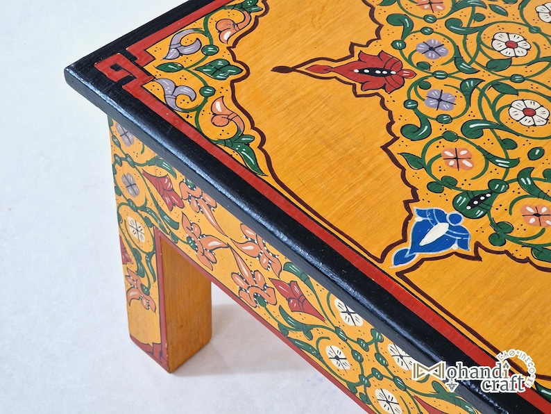 May include: A low, wooden table with a vibrant, hand-painted design. The tabletop is a warm yellow with intricate floral patterns in red, green, white, and blue. The edges are outlined in black and red. The table's legs are a natural wood color.