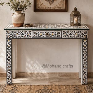 Può includere: Un tavolo console rettangolare caratterizzato da un intricato motivo floreale intarsiato in bianco e nero sul piano, sulla parte anteriore e sulle quattro gambe. Il tavolo include un cassetto centrale con un piccolo pomello rotondo e il testo @Mohandicrafts.