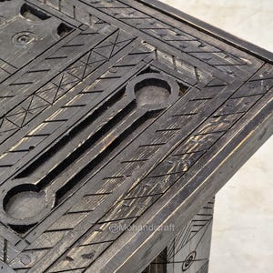 May include: Close-up of a dark wooden table corner with detailed carved geometric patterns. The table has a distressed finish, with visible wood grain and a mix of dark and light tones. The design includes linear and circular elements.