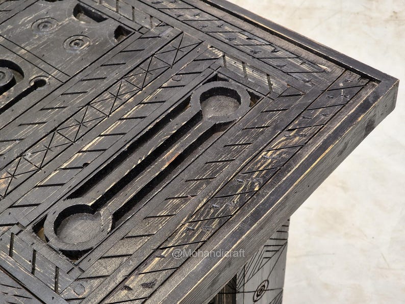May include: Close-up of a dark wooden table corner with detailed carved geometric patterns. The table has a distressed finish, with visible wood grain and a mix of dark and light tones. The design includes linear and circular elements.