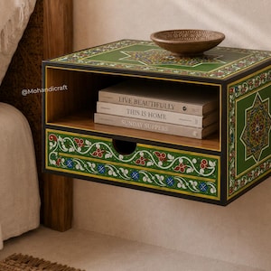 May include: A wall-mounted wooden nightstand featuring a green finish with intricate floral patterns and a central star motif. The unit includes an open shelf holding three books titled Live Beautifully, This Is Home, and Sunday Suppers, plus a drawer.