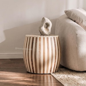 May include: A decorative side table with a striped pattern in shades of brown and cream. A stone sculpture sits atop the table. The table is next to a beige sofa and a patterned rug. The table is approximately 18 inches in diameter and 20 inches tall.