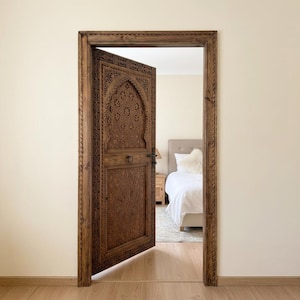 May include: A detailed, open wooden door with intricate carvings, set within a matching frame. The door reveals a glimpse of a bedroom with a bed and neutral-toned decor. The wood has a warm, brown hue.