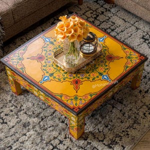 May include: A square, hand-painted coffee table with a vibrant yellow surface and intricate floral patterns in red, blue, and green. The table features a decorative border and is adorned with a vase of orange flowers and a small lantern. The text "@Mohandicraft" is visible.