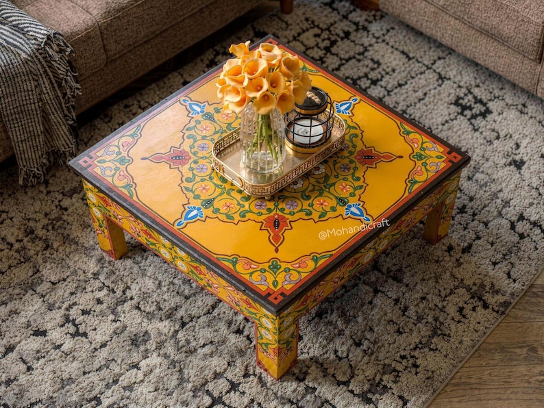 May include: A square, hand-painted coffee table with a vibrant yellow surface and intricate floral patterns in red, blue, and green. The table features a decorative border and is adorned with a vase of orange flowers and a small lantern. The text "@Mohandicraft" is visible.