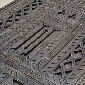 May include: Close-up of a dark grey wooden surface with intricate geometric patterns. The design features repeating diamond, triangle, and linear shapes, creating a textured, carved appearance. The wood has a distressed finish.