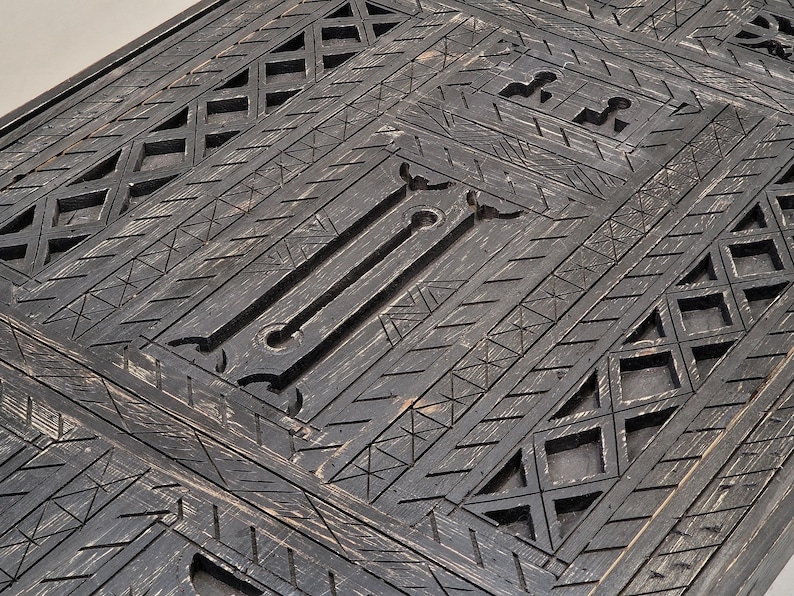 May include: Close-up of a dark grey wooden surface with intricate geometric patterns. The design features repeating diamond, triangle, and linear shapes, creating a textured, carved appearance. The wood has a distressed finish.