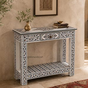 Può includere: Un tavolo console rettangolare caratterizzato da intricati motivi floreali intarsiati in bianco e nero sulla superficie, sulle gambe e sui frontali dei cassetti. Il tavolo include un cassetto centrale singolo con un piccolo pomello e un ripiano inferiore, contrassegnato dal testo Mohandicraft.
