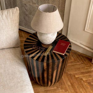 May include: A circular side table with a striped design in dark brown and black. A beige lamp with a white base is on top, alongside a red book and spectacles. The table is beside a beige sofa.