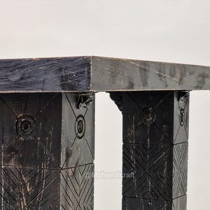 May include: Close-up of a black wooden table with a rectangular top. The table's legs are square and feature carved geometric patterns and circular designs. The table is made of dark wood and has a rustic appearance.