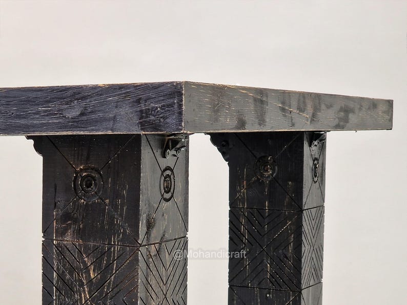 May include: Close-up of a black wooden table with a rectangular top. The table's legs are square and feature carved geometric patterns and circular designs. The table is made of dark wood and has a rustic appearance.