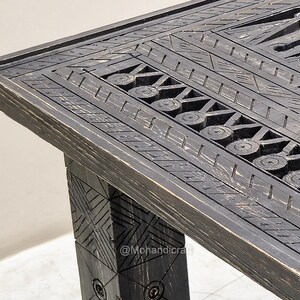 May include: Close-up of a dark stained wooden table with intricate carved geometric patterns. The table has a rectangular top and a sturdy leg, showcasing detailed craftsmanship. The design includes circles and linear elements.