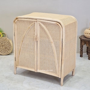 May include: A light-coloured rattan cabinet with two doors and a rounded top. The cabinet has a woven texture and is supported by four short legs. A woven handbag and a small wooden table are in the background.