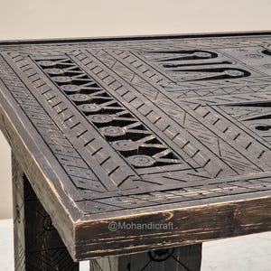 May include: A dark brown wooden table with detailed carved geometric patterns. The table's surface features a design of lines, circles, and shapes, creating a textured appearance. The table's legs are visible, matching the dark brown colour and carved design.